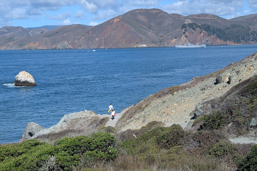 in the foreground: a trail winds through rocky cliffs, dotted with shrubs. Beyond, a blue bay. Beyond that, the marin headlands, mostly brown but dotted with green