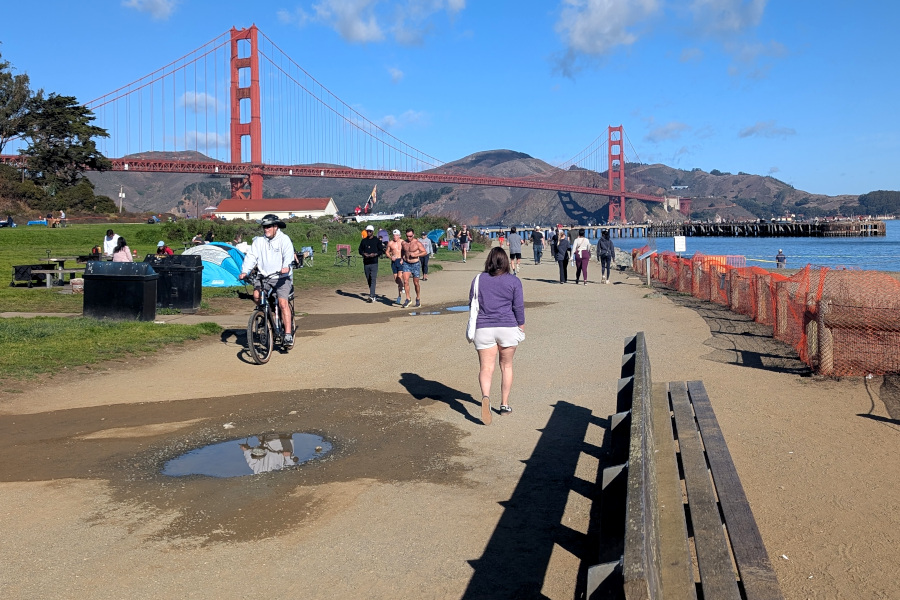 in the foreground, walking path by the bay with walkers and
		joggers. In the background, the Golden Gate Bridge