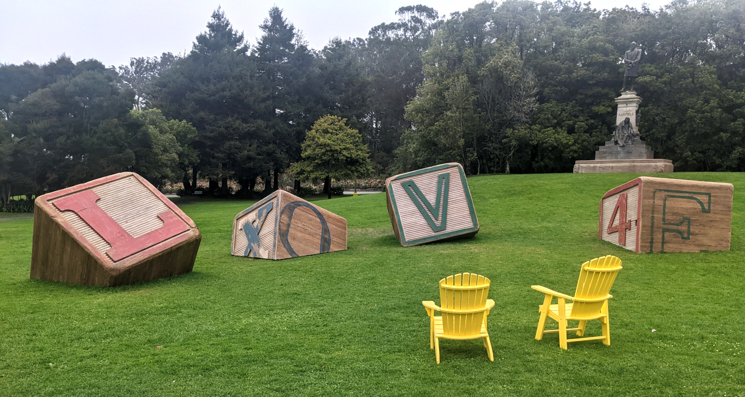 [picture of art installation that looks like four giant alphabet blocks embedded in lawn. From this side, the blocks' letters spell out 'Love' or maybe 'Lqve', the O seems off-center]