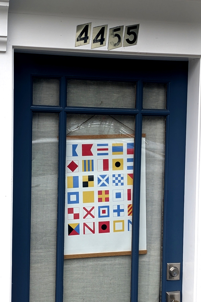 [door, much of which is glass; behind the glass hangs a sign; the sign shows the maritime signal flag alphabet plus four flags that kinda look like the letters in 'noon']