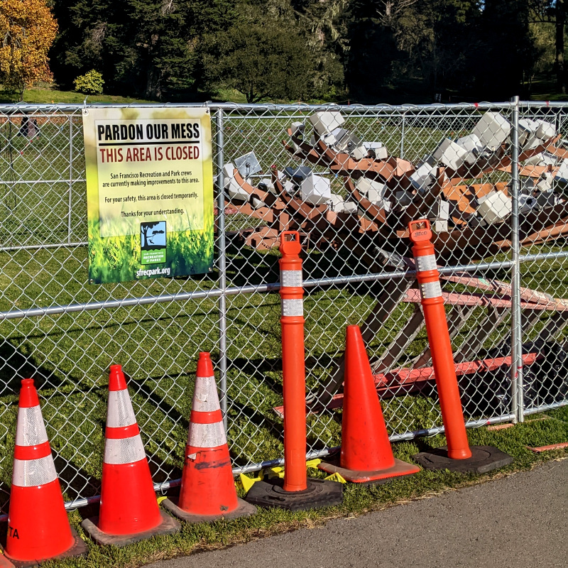[photo: behind a chain link fence and traffic cones, a pile of plasticky-looking dendrite forms to be assembled into some light-up trees. A sign reads "pardon our mess"]