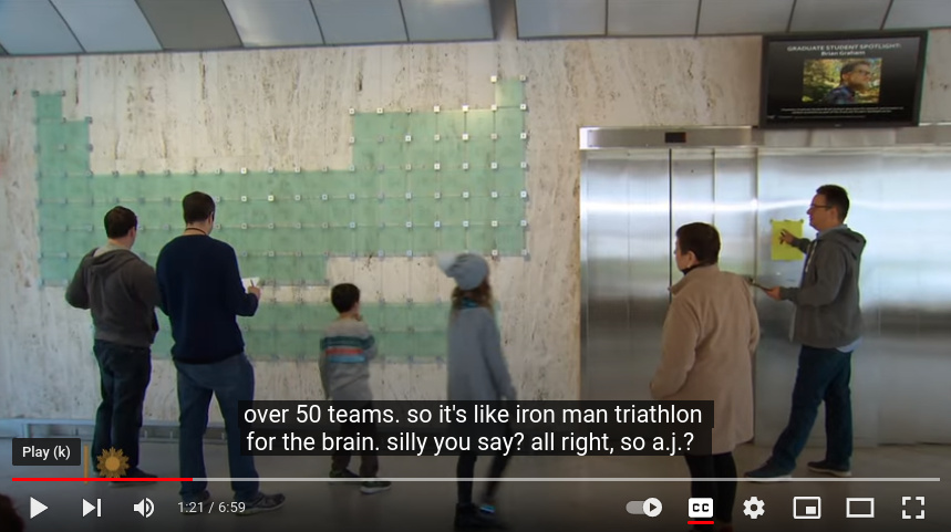 still frame from video. some nerds stand before a large periodic table of elements. Closed caption reads: over 50 teams so it's like iron man triathlon for the brain. silly, you say? all right, so a.j.