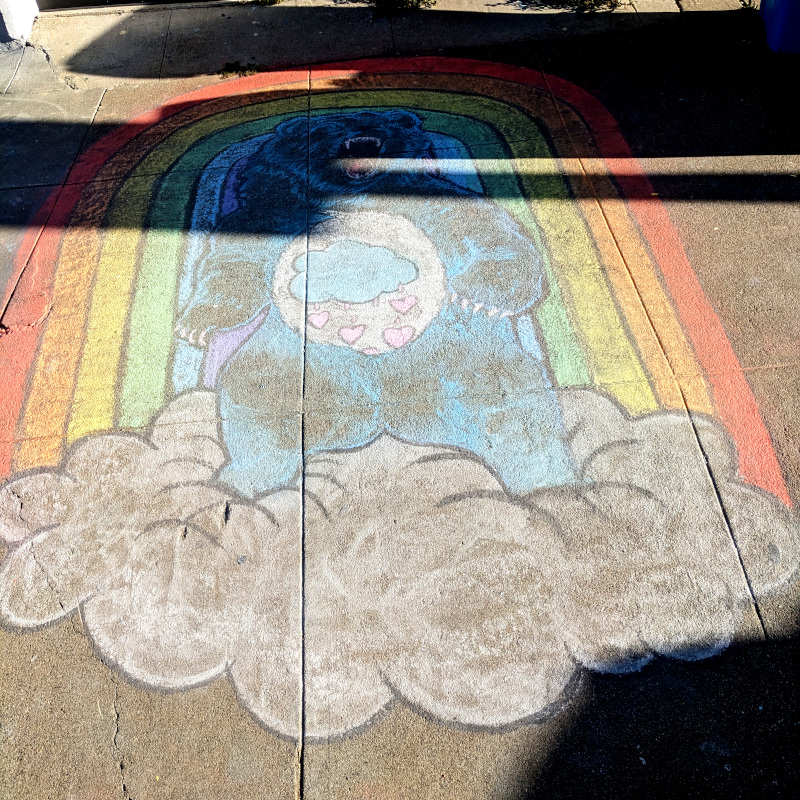 Sidewalk chalk art of blue bear with stormy heart chest decoration against a rainbow background