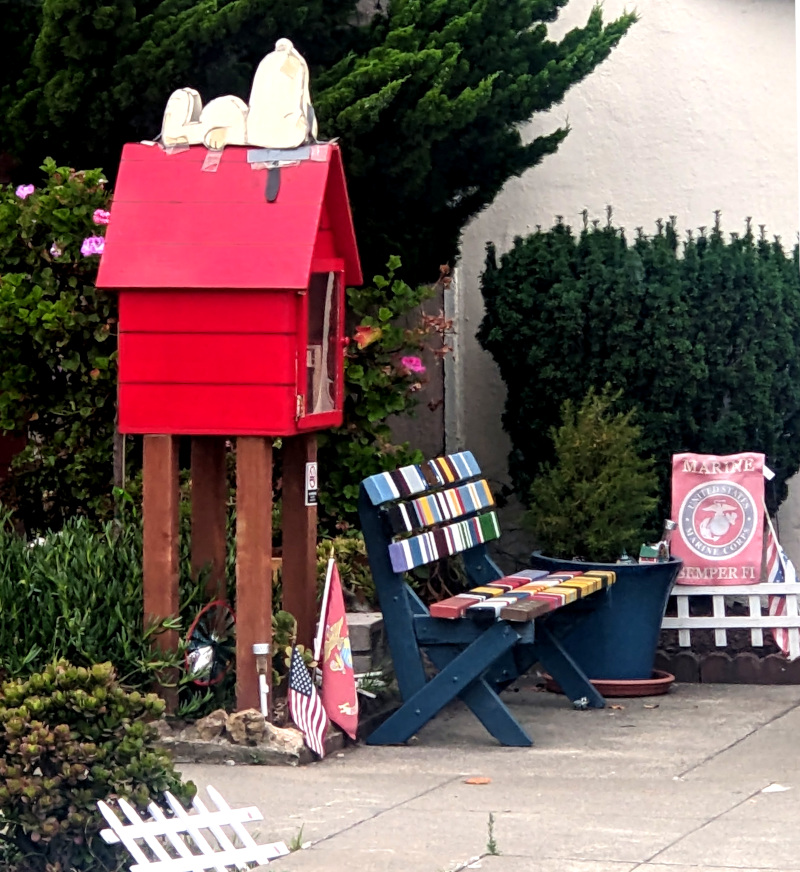 A little free library in the form of a red doghouse with Snoopy the dog napping atop it. Also a bench and some USMC decoration