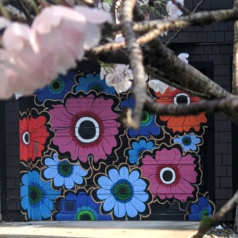 photo of flowers. In foreground, out of focus, some cherry blossoms. In background, in focus, a garage door mural depicting flowers, maybe poppies? I dunno, I'm not a big flower guy
