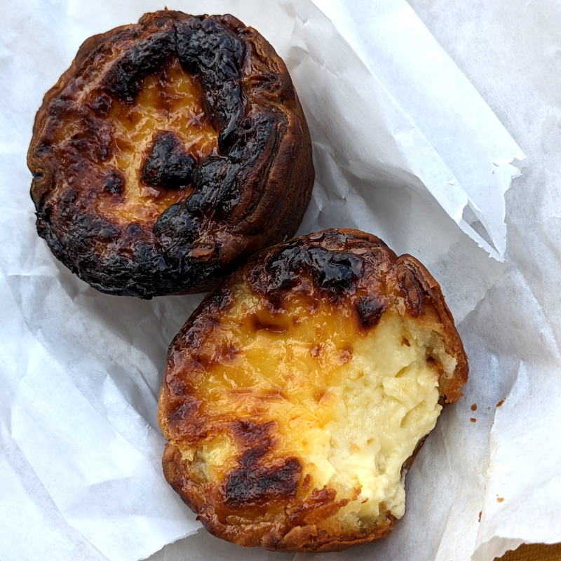 two pastries (one with a bite taken out of it) sit on the little bags they came in. They look like regular egg custard tarts, except carmelized on top like a flan and their crusts look croissant-darkish instead of pie-lightish