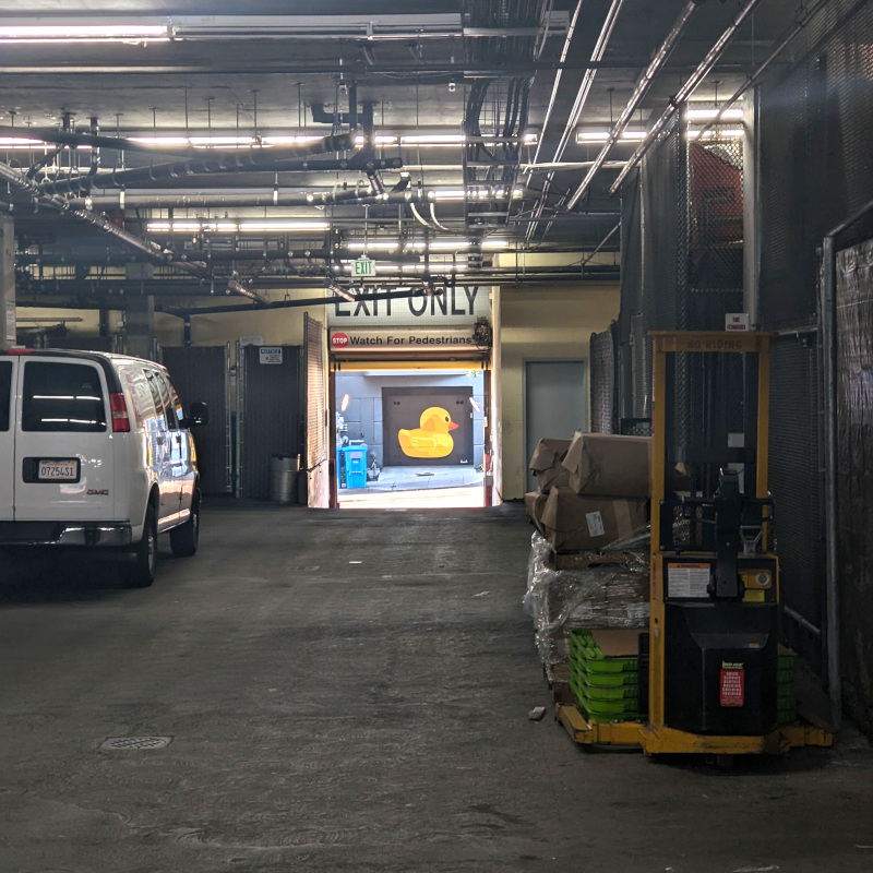 a parking garage. there's a forklift and an exit door. out the exit door, there's a stencilly-looking rubber duck mural