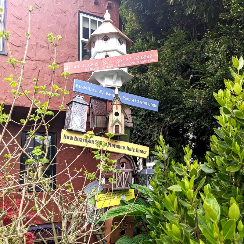pole to which is affixed decorated birdhouses and signs. One sign says 37 degrees, 43 minutes, 47.6832 seconds N 122 degrees 27 minutes 53.6292 seconds [implied West] Mt Davidson Manor. We need to know this. Another sign says Birdville's #1 Realtor, Paul 415-902-8969. Another sign says Now boarding SFO to Florence Italy, direct