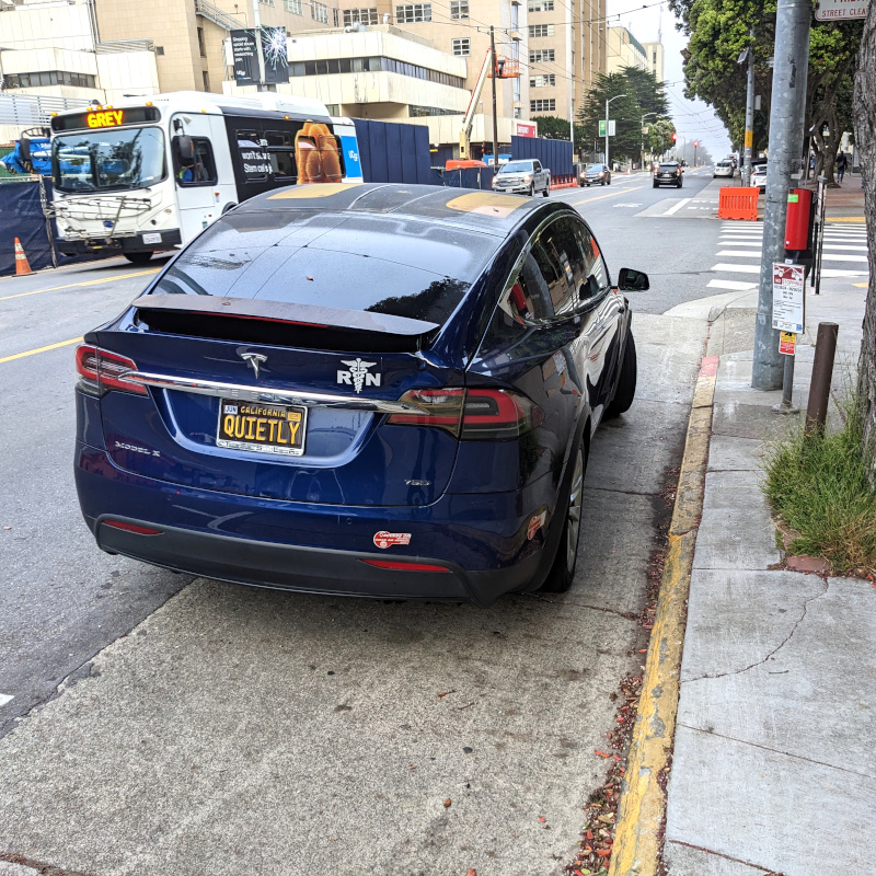 photo of parked car, seen from the rear. It has a "R☤N" bumper sticker. The (California) vanity license plate reads "quietly". The car is a Tesla model X