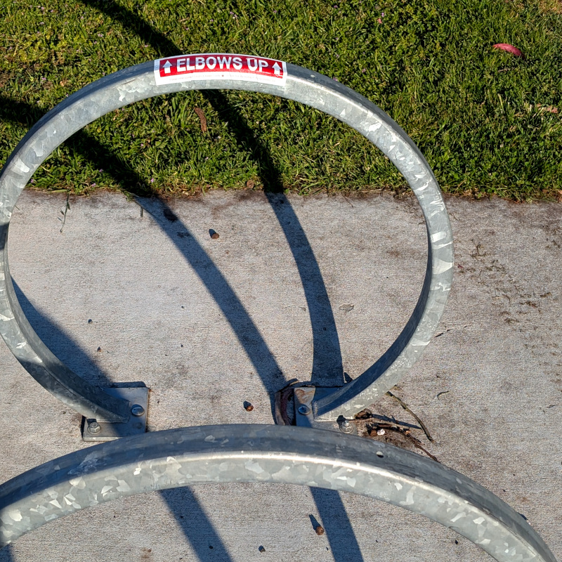 Bike racks, shadows. One rack has a sticker: "↑ Elbows Up ↑"