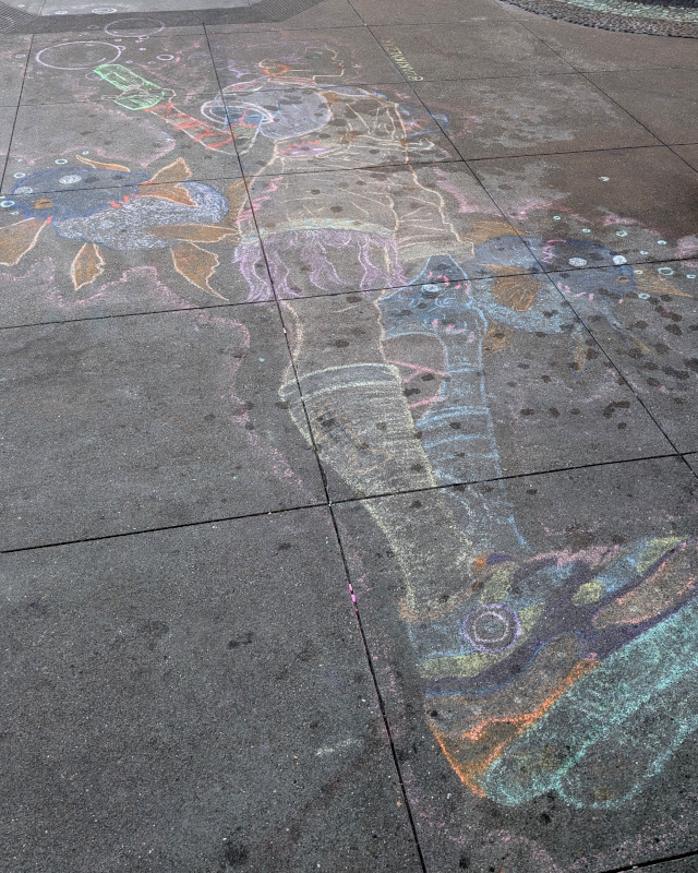 sidewalk chalk art. a human-ish figure floats above us, flanked by giant goldfish. the goldfish exude bubbles. the figure wields a handgun that emits bubbles, an air gun?