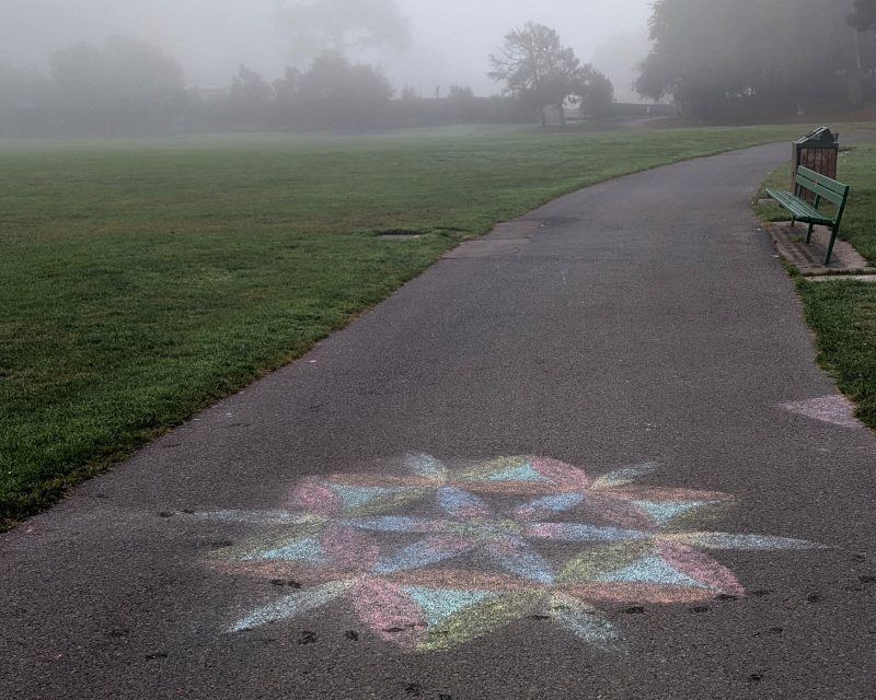 in the foreground, a sidewalk wiggles between a field and a grassy hill. drawn on the sidewalk in colored chalk are some circles and arcs which form a hexagonal construction suggesting triangles. in the background, trees obscured by thick fog. really thick fog. would you believe me if i told you there's a dog and a dog-walker back there?