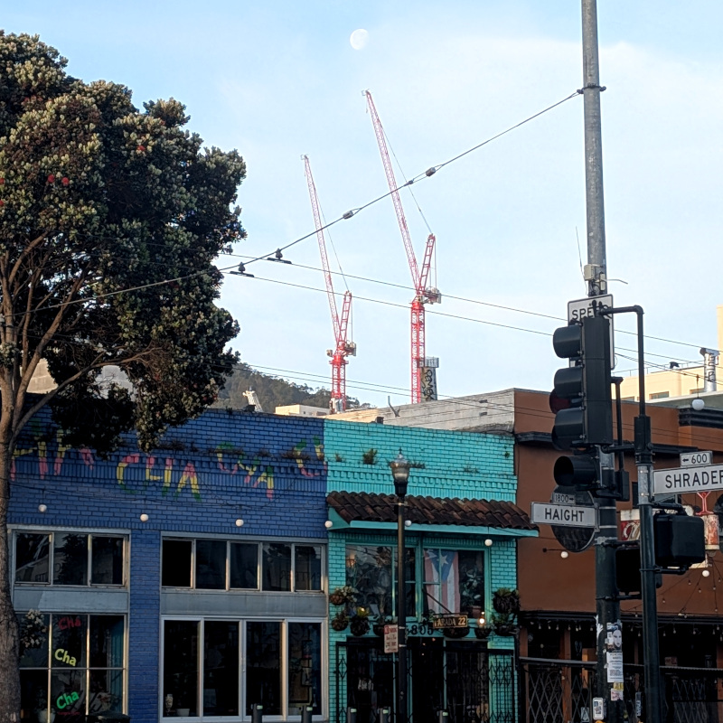 in the foreground, cropped view of the intersection of Haight and Shrader streets. You can see the restaurant Cha Cha Cha, a couple of its neighbors, and street signs. In the background, behind the buildings, are a couple of orange cranes building the new hospital at UCSF Parnassus. Above one of the cranes is a faint white circle; it's the moon, I promise