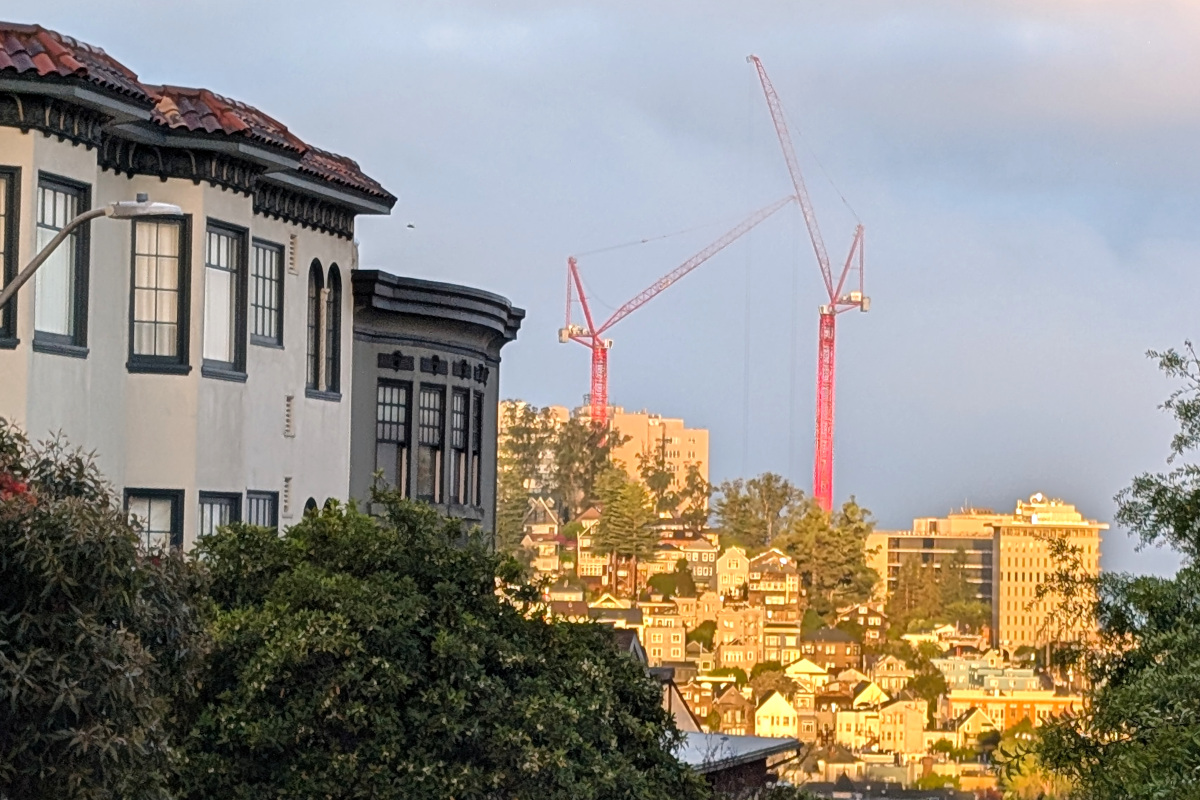 a couple of far-away big orange construction cranes loom over the new UCSF Parnassus hospital going up. They're framed by some buildings and trees that are closer, this photo having been taken from a hilly street several blocks away