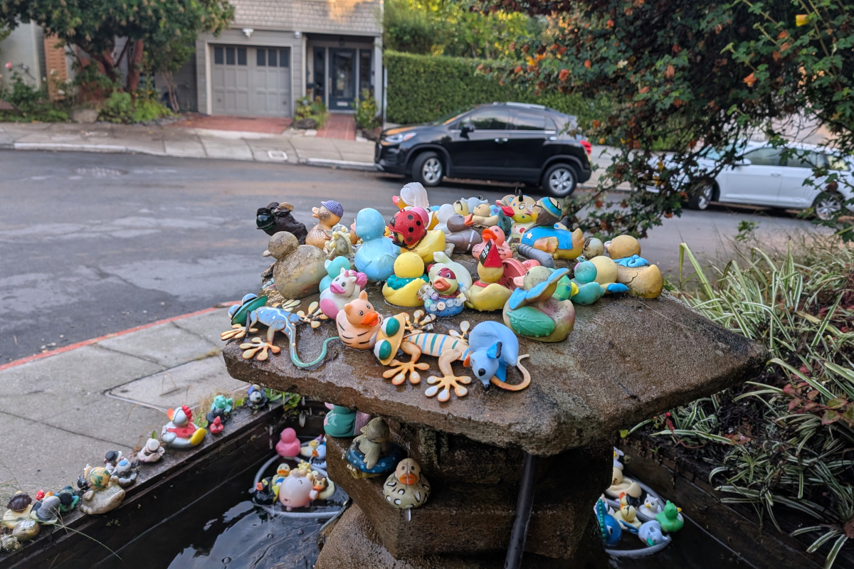 a flotilla of rubber duckies and similar plastic bath toys sits atop a fountain in the form of a sort of Japanese stone lantern maybe? I'm not a garden decor expert