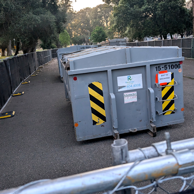 some dumpsters surrounded by a temporary fence; some trees loom in the background. A piece of paper taped to the closest dumpster reads: Bay to Breakers