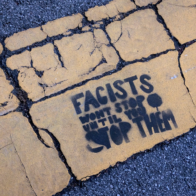 stenciled graffito on yellow crosswalk paint on asphalt. It reads: Facists wont stop until you [picture of crossing guard's handheld stop sign] stop them