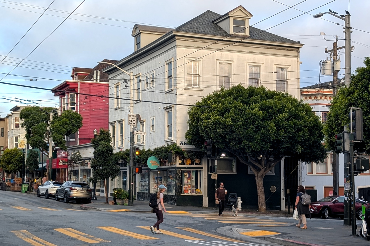 that t-shirt shop at the corner of Haight/Ashbury in morning light. closer to the camera, some tourists mosey around, realizing they've arrived at this tourist site too early to see hustle and/or bustle