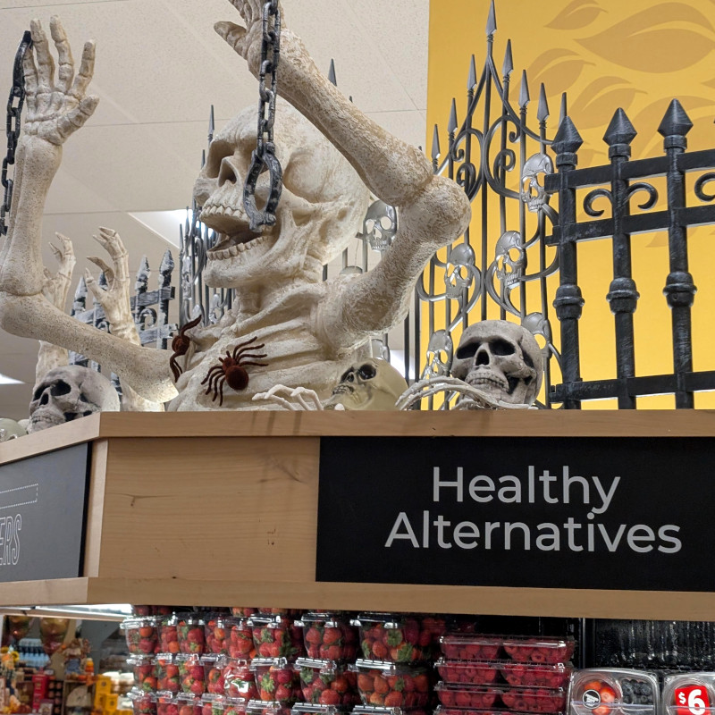 photo of supermarket interior. at the top, a Hallowe'en display features skeletons and skulls. The display sits atop a cabinet labeled 'Healthy Alternatives' containing strawberries