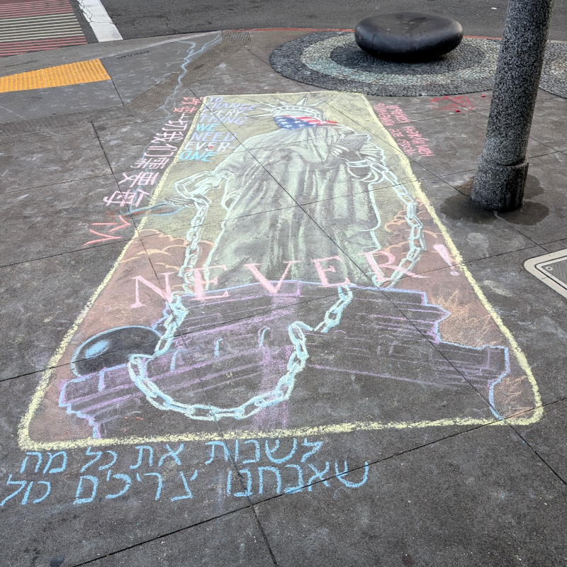 Sidewalk chalk art. We are looking up at an altered Statue of Liberty against warm-colored clouds. Altered: Instead of broken chains of freed slaves at her feet, her wrists are cuffed to chains. She wears the US flag as a bandanna. There is big text superimposed, all caps: Never! In the upper-left corner, more text: To change everything we need everone (sic, dangit). Around the border is text in languages I cannot read, alas; I guess Chinese, Hebrew and Greek-or-Russian?