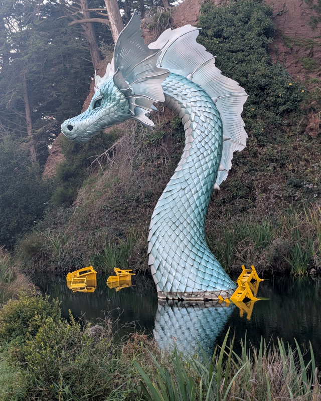 statue of a sea serpent rises from a pond, surrounded by several half-submerged bright yellow Adirondack chairs