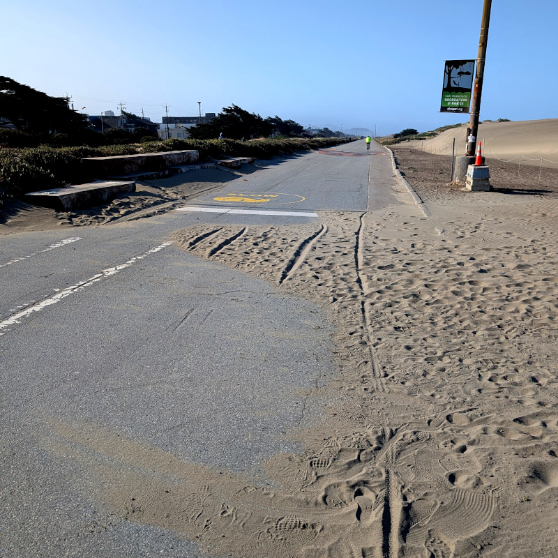 Stretch of road surrounded by dunes. Some sand has covered part of the road