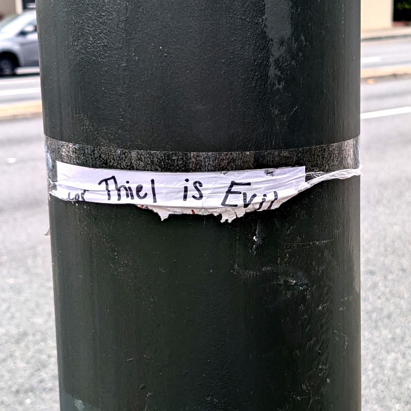 a mostly-torn-down flyer on a utility pole. The scrap of paper reads: (Pe)ter Thiel is Evil