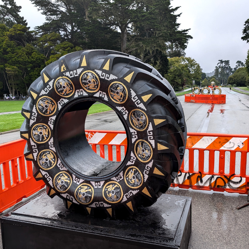 in the foreground, a big vehicle tire with gold-colored art painted on it. In the background, a set of three tires, also with gold paint. Way further back in the background, a sort of golden sphere