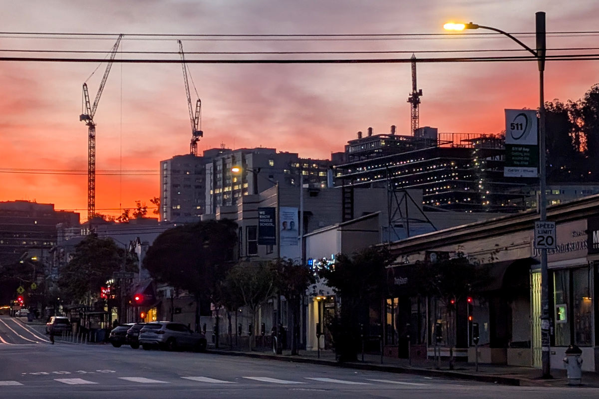 in the foreground, a retail street. behind, dimly-lit big buildings on a hill; when you peer more closely, you notice the one on the right is under construction. above the buildings hover some cranes, confirming that construction's going on. in the very background, colors of sunrise