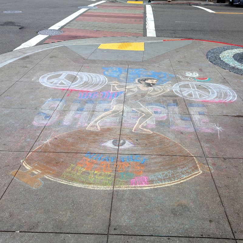faded sidewalk chalk art. There's a lot going on here. Text in the background: We the sheeple. Text at the bottom: What they hate in you is missing in them. Depicted: A weightlifter lifts a barbell. He's standing on an upside-down wok that has one eye (which is looking up at the weightlifter). The weights on the barbell are peace signs.