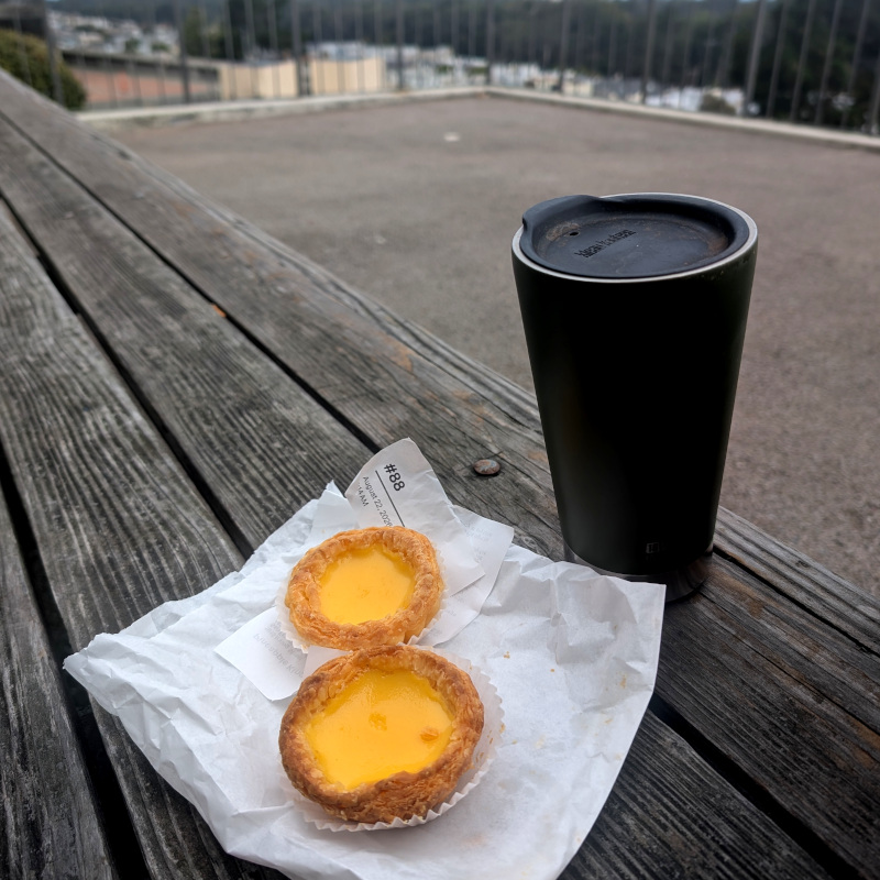 some food on a bench outside. there's a sippy-cup with coffee next to it. the food: a couple of 'dan tats' 蛋撻 egg custard tarts w/ a receipt saying mine was order #88