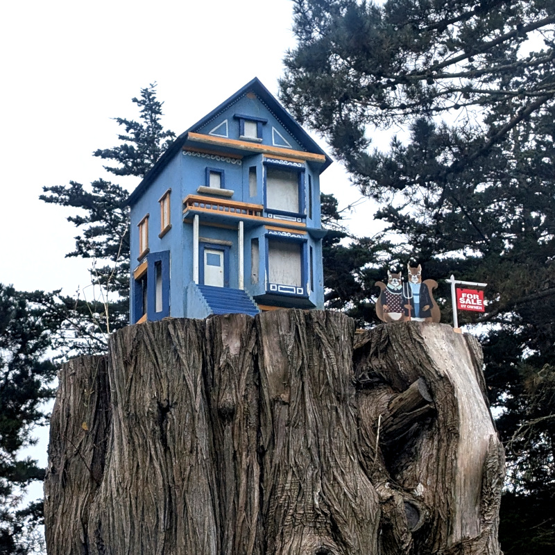 tree stump atop which sits a model house. by the house is an American Gothic squirrel couple. Beside them, a little real estate sign that says 'for sale by owner'