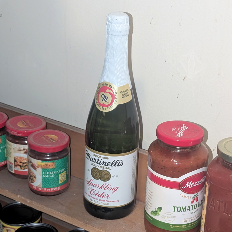 kinda dusty shelf with chili garlic sauce, pasta sauce, and featuring a bottle of sparkling apple cider kept on hand in case of sudden celebration occasion