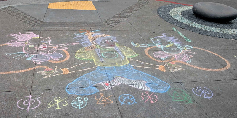 sidewalk chalk art: person dressed in a t-shirt with the Lucca Deli logo sits in lotus position. Their hands hold small spheres from which emerge hoses which seem to anchor a couple of small demons. Below are eight mysterious symbols