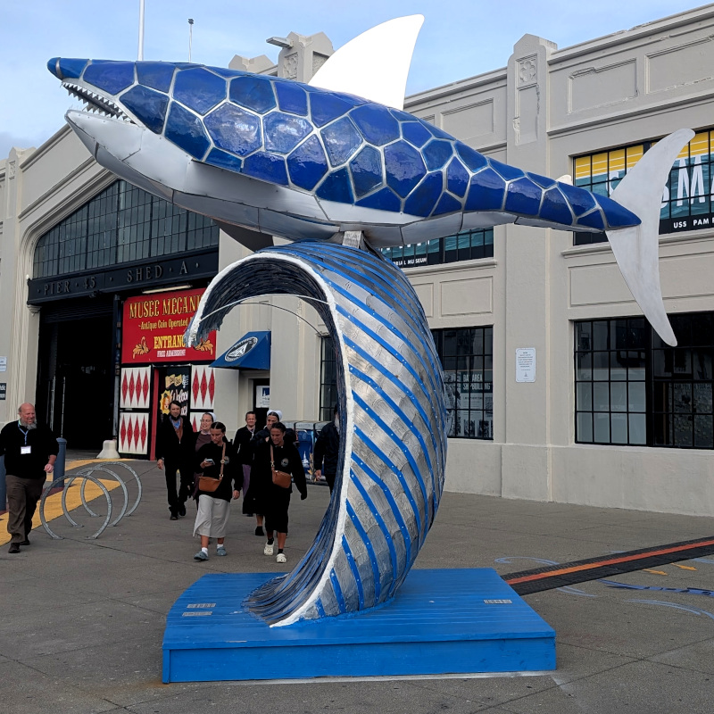 Statue of a shark belly-surfing a wave. it's gray, but the top looks like someone put a mosaic-filter blue patch on top.