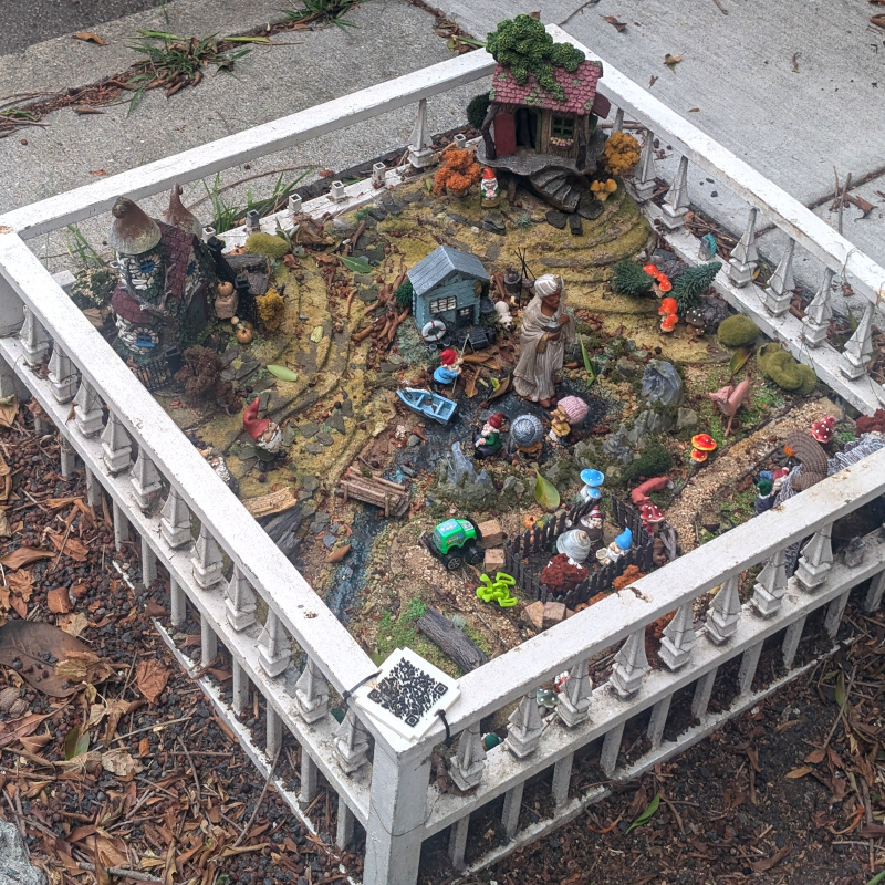 hole in the sidewalk sized such that you might expect a tree. instead, there is a variety of toys making a sort of fantastic village scene. what kind of toys? uh, houses, gnomes, big guy in a turban, mushrooms, and many many more