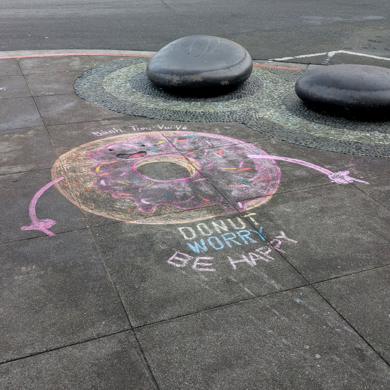 Sidewalk chalk art: an anthropomorphic donut smiles at you in an arguably-appetizing manner, making the finger-guns gesture. Text above: Bánh Tiêu Vui Vé. (Please don't throw rocks at me if I got those accent marks wrong; I tried my best.) Text below, all caps: Donut Worry, Be Happy