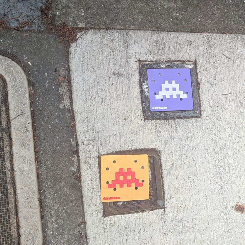 patch of sidewalk with a couple of covered drains. instead of the usual plain drain covers, these are brightly colored with raised pixel art reminiscent of the old Space Invaders game. They are signed @draINvader