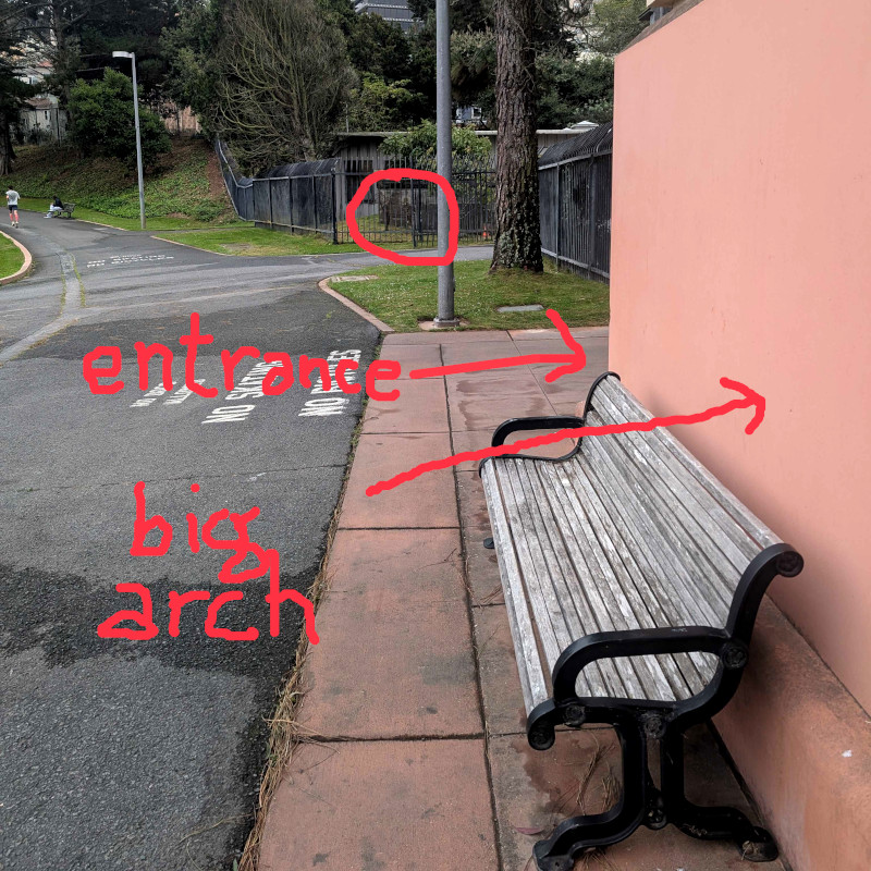 altered photo: path, lawn, fence, hedge, the edge of a big arch, and an ornament-thing. the alteration: someone has circled the ornament-thing and also written 'big arch' with an arrow pointing to the edge of the big arch plus 'entrance' with an arrow showing where the entrance is, next to the arch