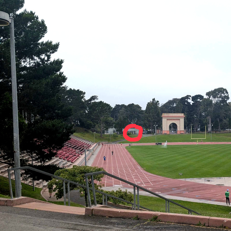 altered photo: view across a running track+soccer field. on the other side of the track is coral-colored big arch. there's also a fence. the alteration: someone has circled a bit of fence in red