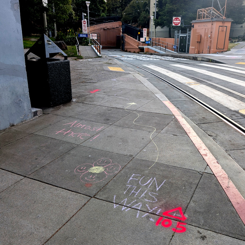 sidewalk chalk message promises "fun this way ↑" pointing down rail tracks that disappear around a bend into a hillside