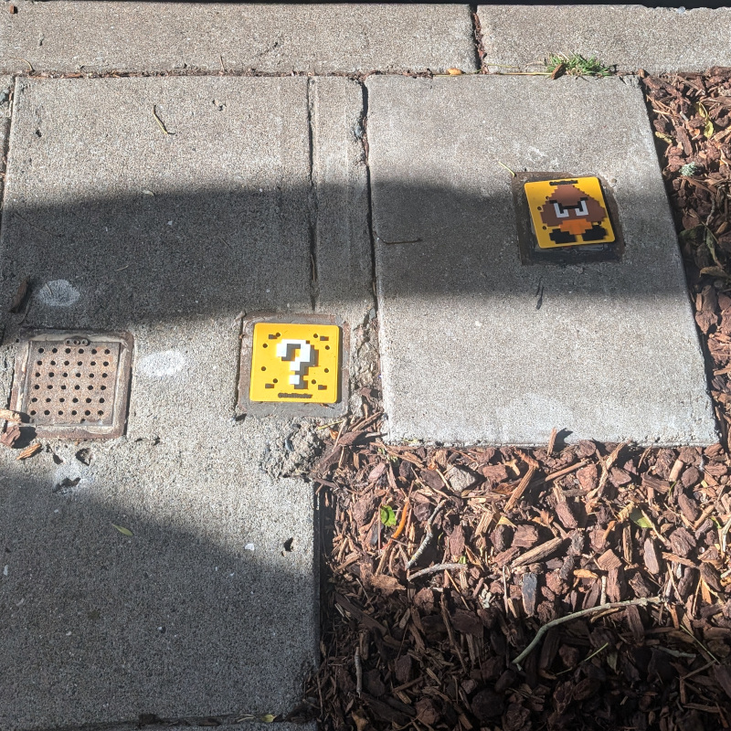 patch of sidewalk with some sewer covers. they're usually made of metal and often stolen. a couple of metal ones have been replaced by ?plastic? decorated to look like a Mario Goomba and question mark