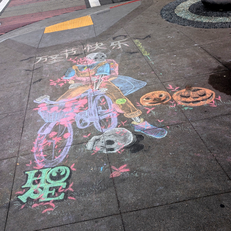 sidewalk chalk art: two ghosts share a bicycle. bats flutter in the background. there are a couple of jack-o-lanterns on the ground, a skull, and a 'hope' with a Star Wars rebellion logo in the O. Also some Chinese that I, alas, cannot read