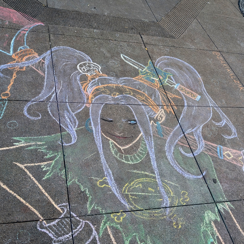 sidewalk chalk art: zooming in on the demon hunter's head. We now see that one hair scrunchie is actually a little skull. The other hair scrunchie is a small demon head with a small sword going through it. That Lucca Deli logo has crossbones behind it. (I assure you I have eaten there and there was no poison in my food; I believe this crossbones is meant to evoke pirate-level ferocity, not poison warning labels)