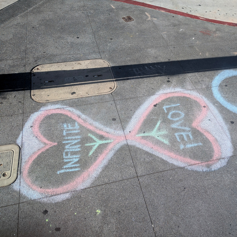 sidewalk chalk art: two hearts placed point-to-point to suggest a ∞ infinity lemniscate symbol. Within one heart is written Infinite; in the other, Love