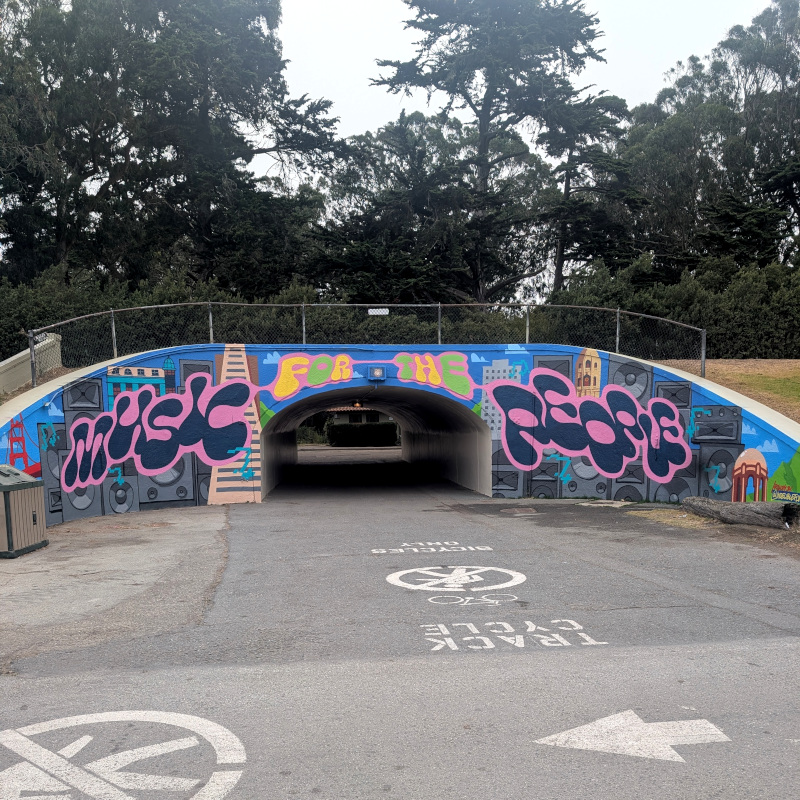 graffiti-style mural around a tunnel mouth. Has small simplified stylized SF landmark buildings side-by-side with giant stacks of speakers. Has text: Music for the People