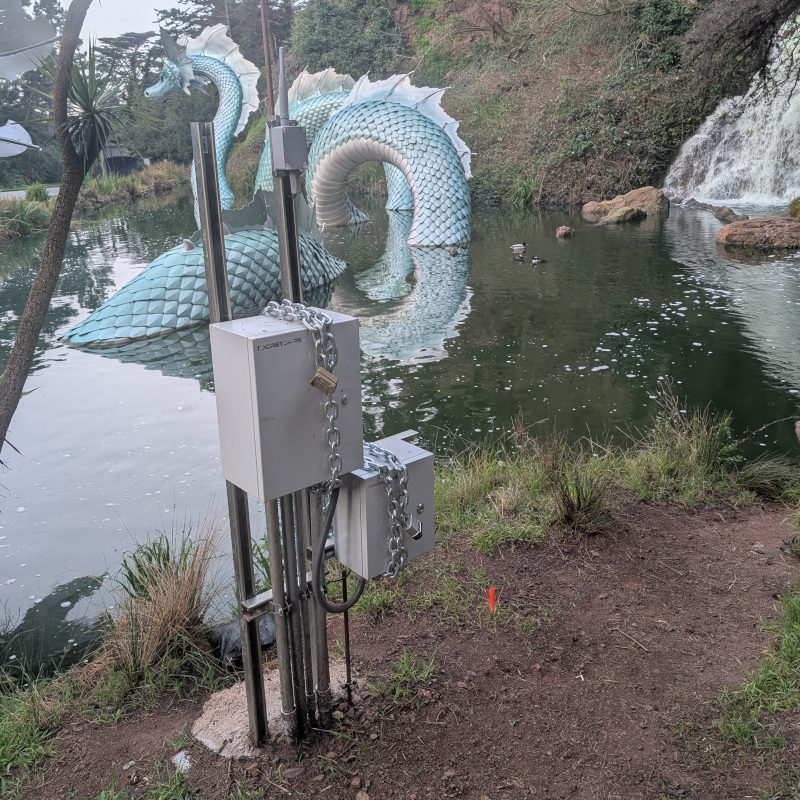 In the background: statue of a sea serpent in a pond; a smidgeon of waterfall. In the foreground: a couple of control boxes mounted on poles. There's also an antenna. Each control box is wrapped in heavy chain, each chain has a choose-your-combination lock