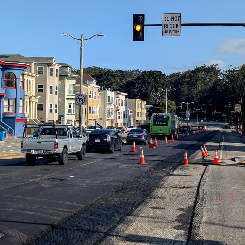 street, normally a few lanes in each direction, but today traffic cones reduce it to one lane each; there is fresh asphalt, still soft enough to stick to your shoe if you walk on it, as it turns out