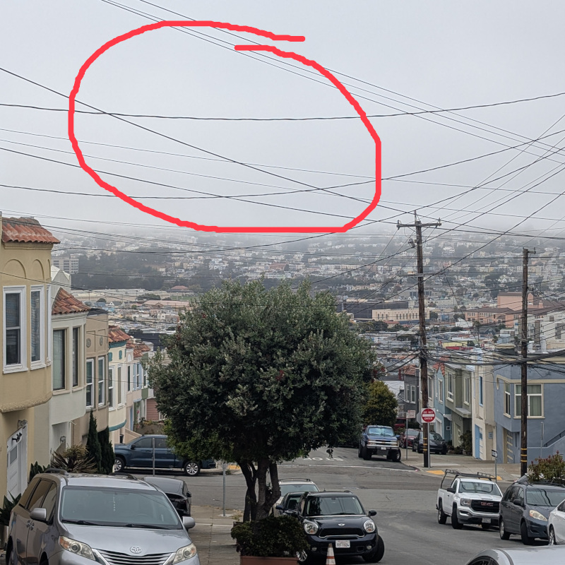 edited photo: view of a street. Above the street is an opaque wall of fog. The photo has been edited: an area of the fog is circled in red, the author's best guess as to Mt. Davidson's location