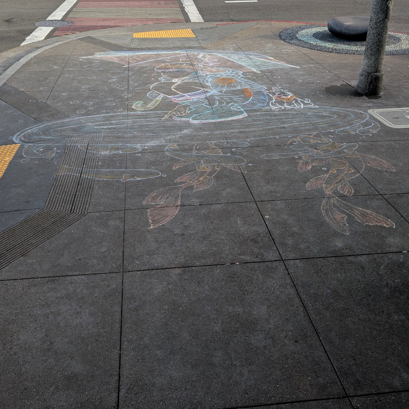 sidewalk chalk art: a lady crouches at the edge of a small pond. A puppy sits behind her. Fish swim below the surface of the pond